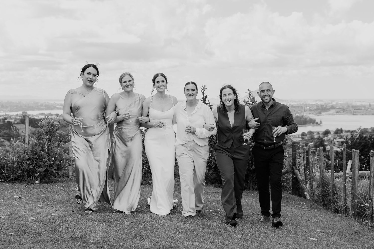 Black and white photo of a bride with bridesmaids and guests smiling outdoors at a scenic wedding venue.