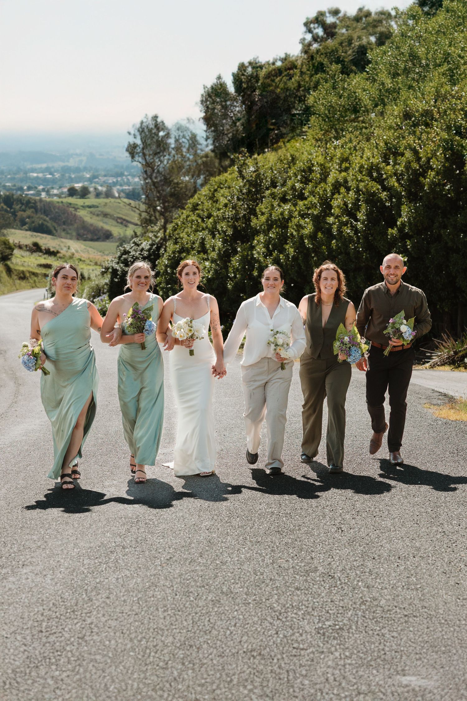 A bride in white walks with bridesmaids in sage green and guests on a scenic road with rolling hills.