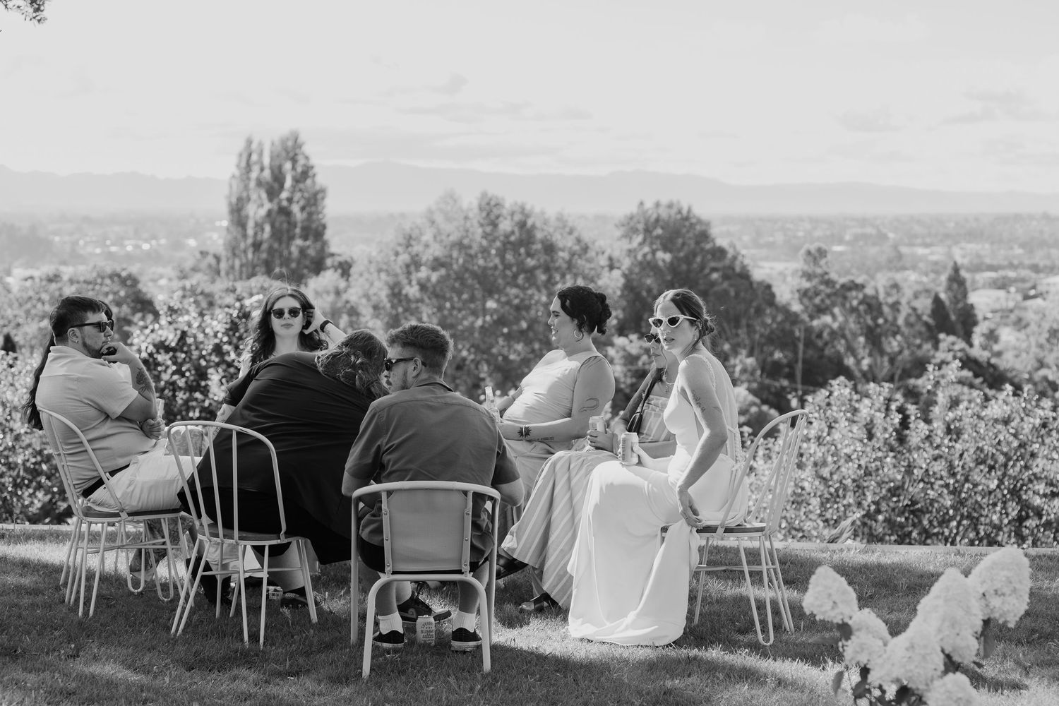 Black and white photo of wedding guests seated outdoors on chairs with scenic landscape views in the background.