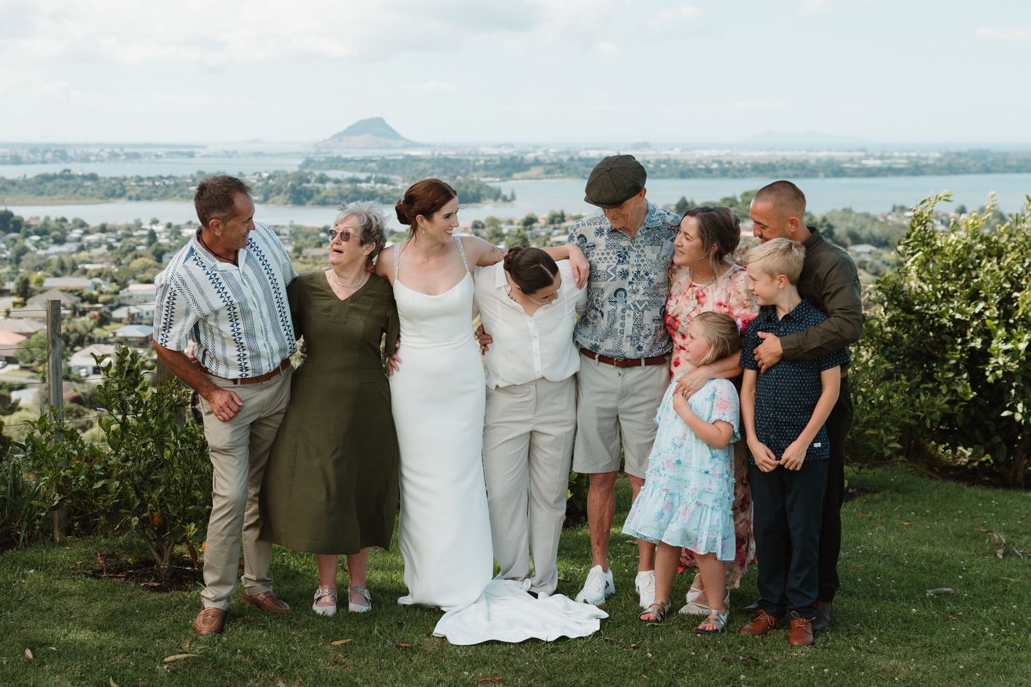 Wedding couple poses with family outdoors, scenic coastal town and hill visible in the background.