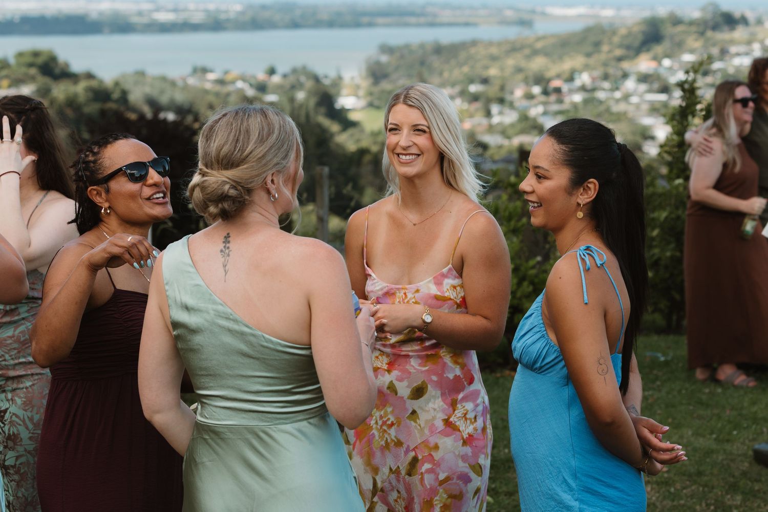 Women in colorful dresses laughing and socializing outdoors at a wedding reception with scenic harbor views.
