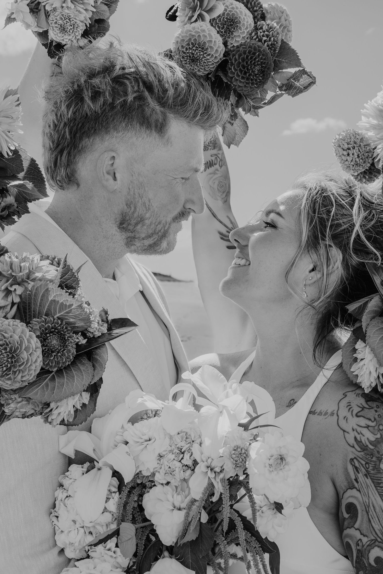 Black and white photo of a smiling bride and groom surrounded by dahlia flowers on their wedding day.
