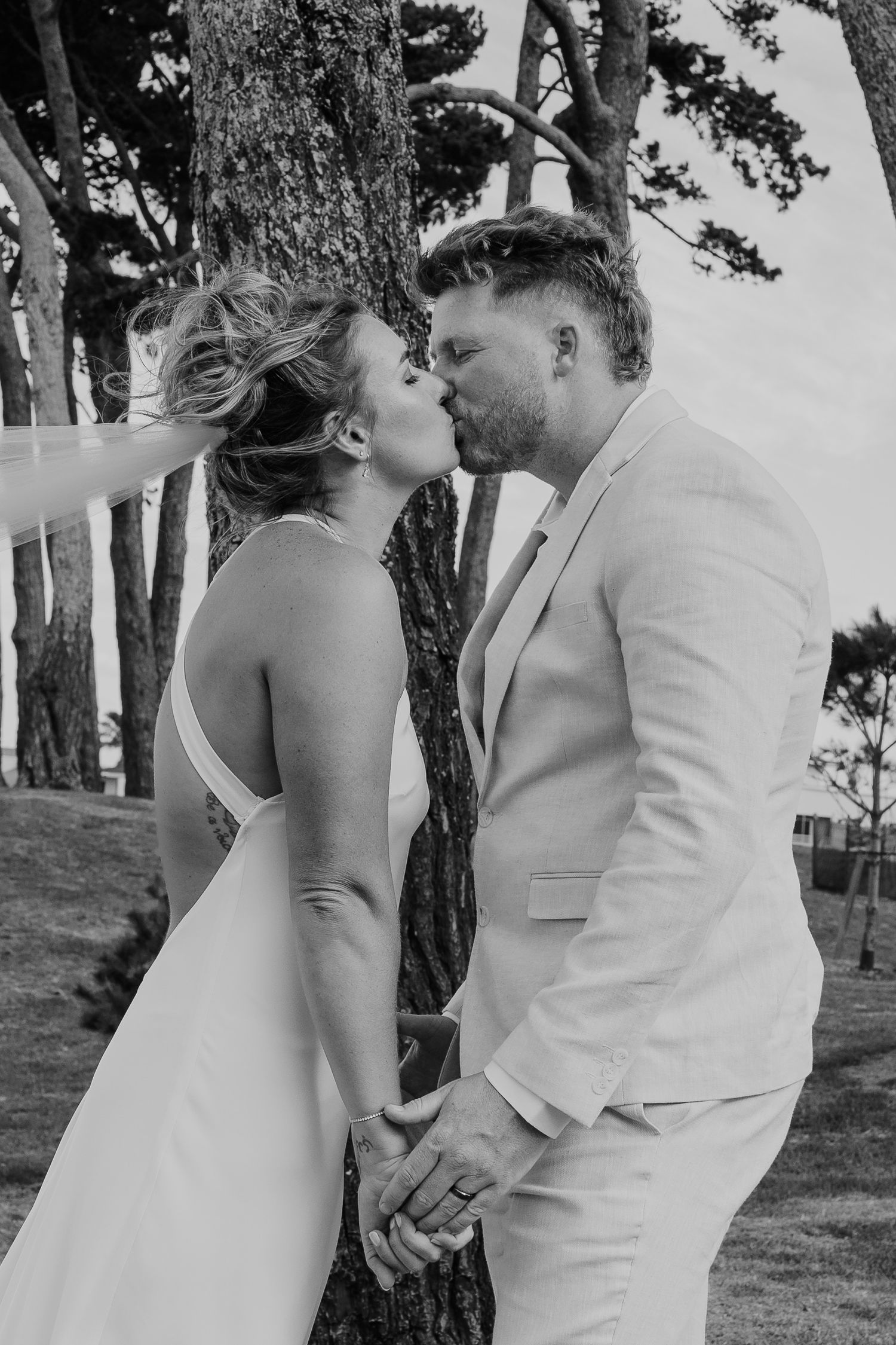 Black and white photo of a bride and groom kissing while holding hands outdoors near trees on their wedding day.