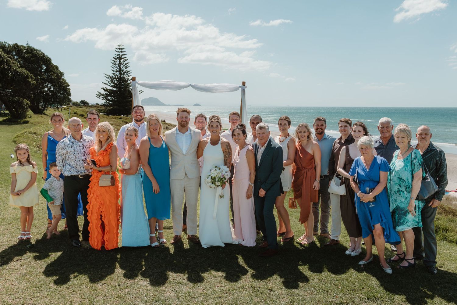 Wedding party group photo on a scenic beachside lawn with ocean views on a sunny day.