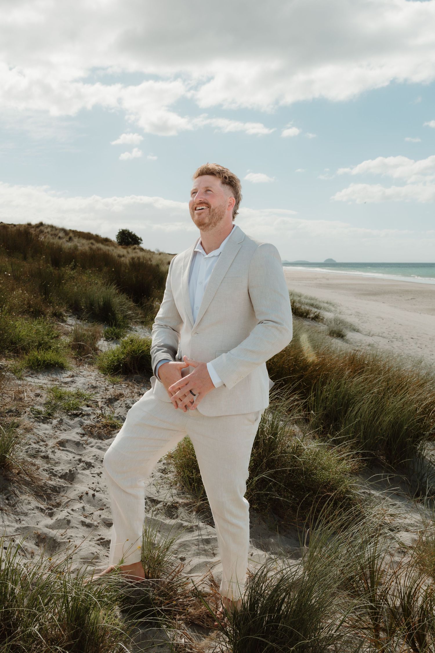 Smiling groom in a beige linen suit standing on sandy beach dunes with ocean views on his wedding day.