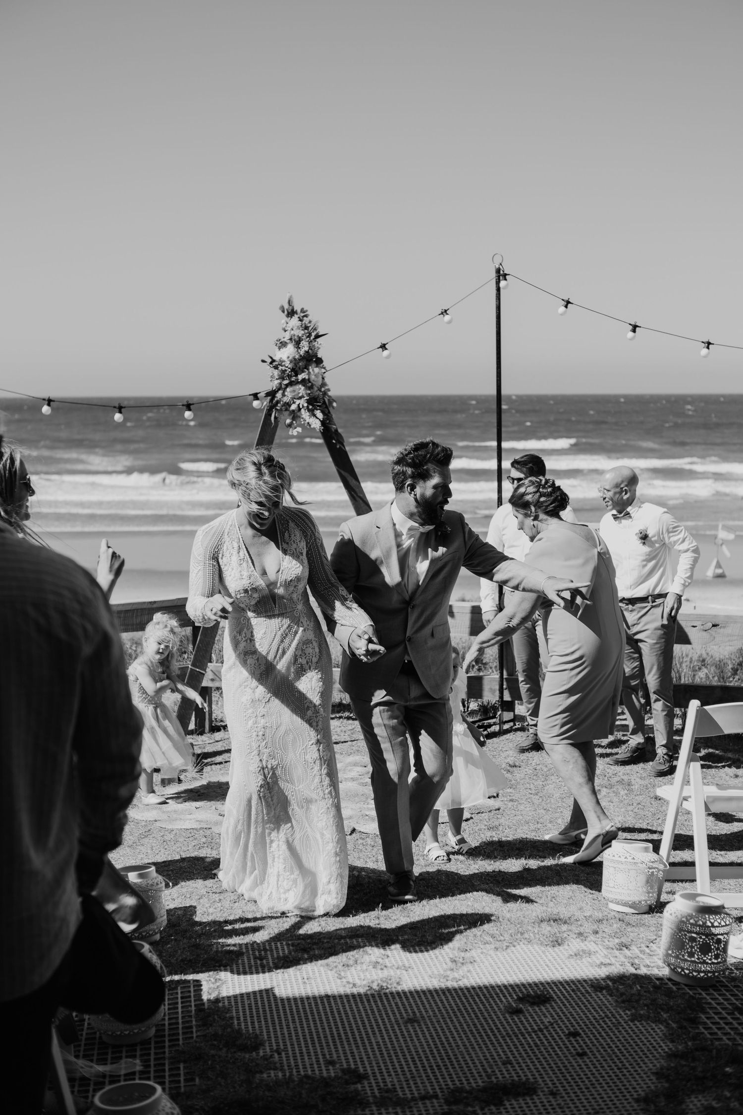 Wedding party celebrates joyfully at a beachside venue under string lights with ocean waves in the background. Papamoa Beach Wedding