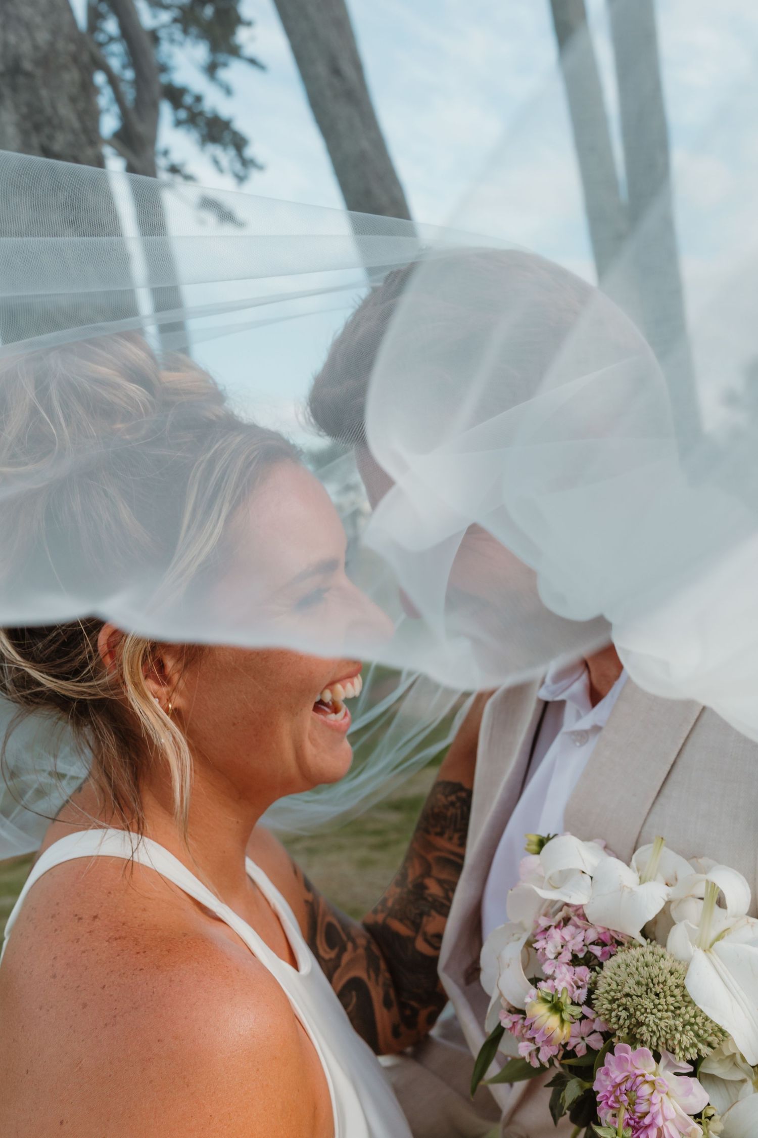 Bride and groom sharing a romantic kiss under a flowing veil, bride holding a pastel floral bouquet outdoors.