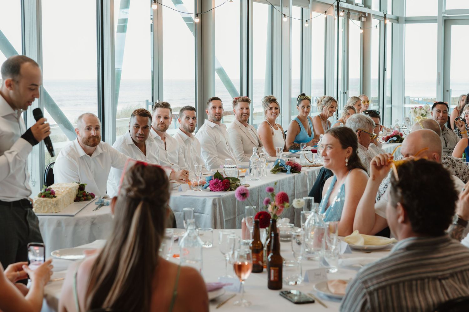 Wedding reception guests gathered at elegant tables while a man gives a toast with a microphone near large windows.
