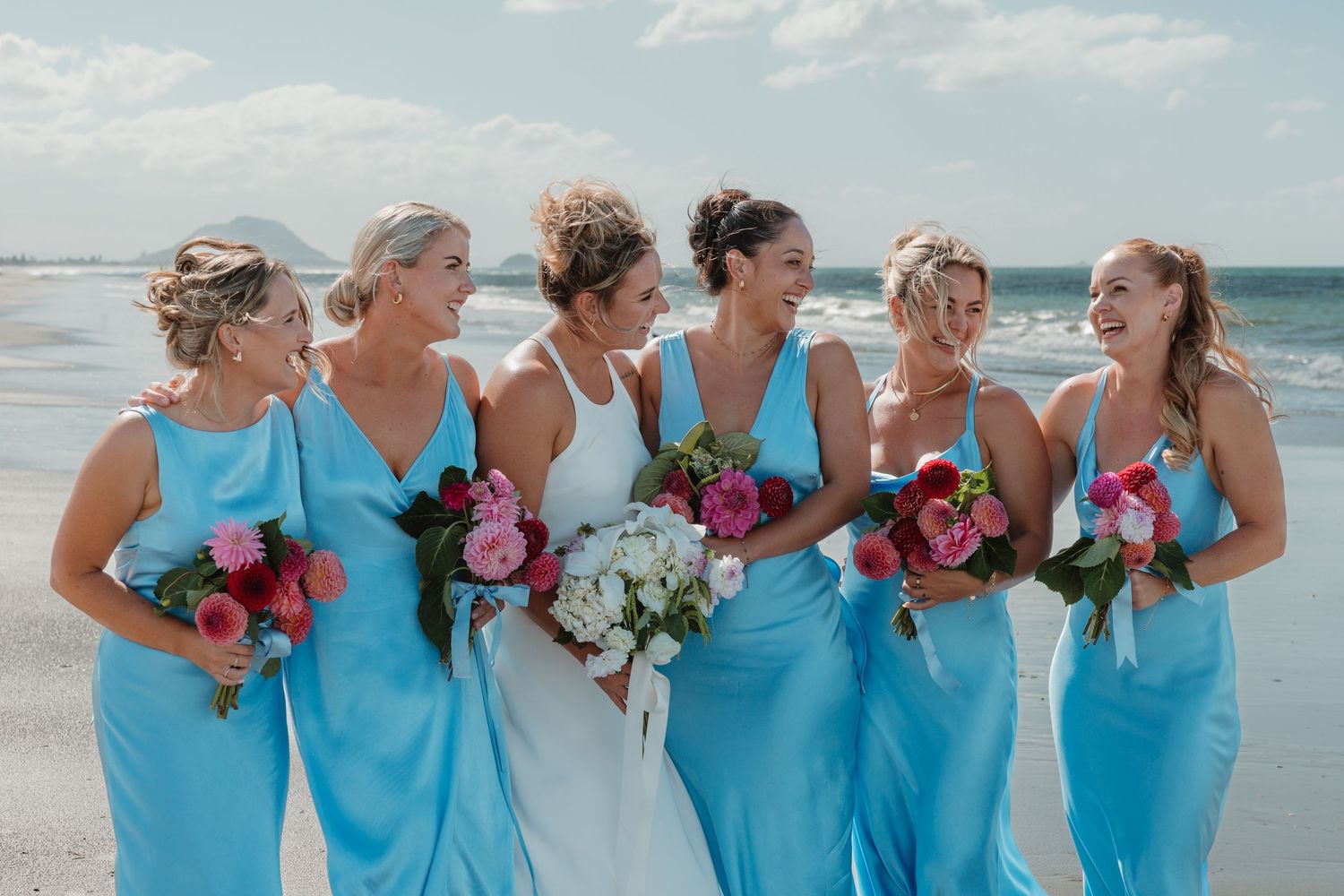Bride in white gown with five bridesmaids in light blue dresses holding colorful bouquets on a beach.