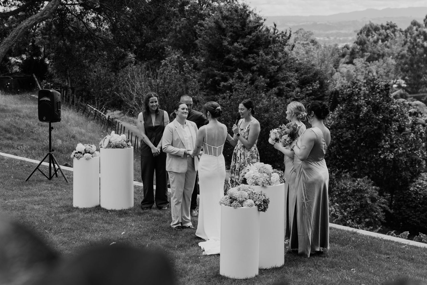 Black and white photo of wedding guests gathered around floral arrangements on white pedestals at an outdoor ceremony.