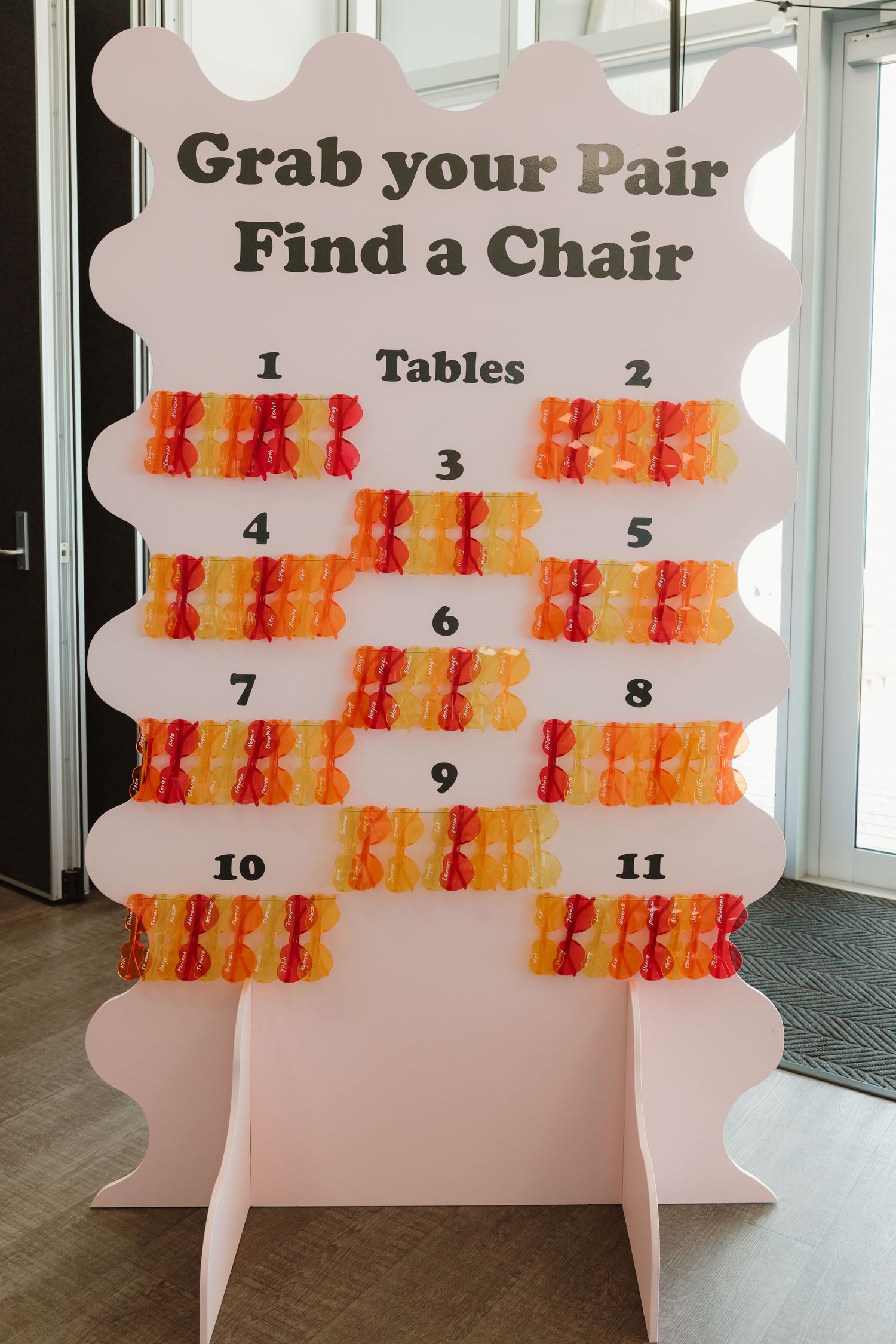 Pink seating chart board with colorful sunglasses, reading Grab your Pair Find a Chair, numbered tables 1-11.