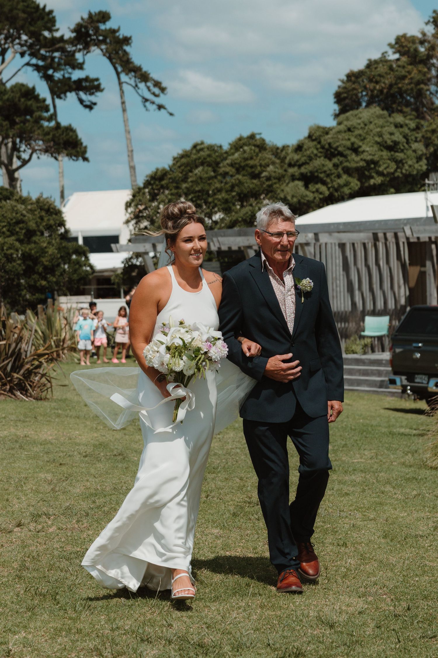 Smiling bride holding white bouquet walks with older gentleman in navy suit at outdoor wedding.