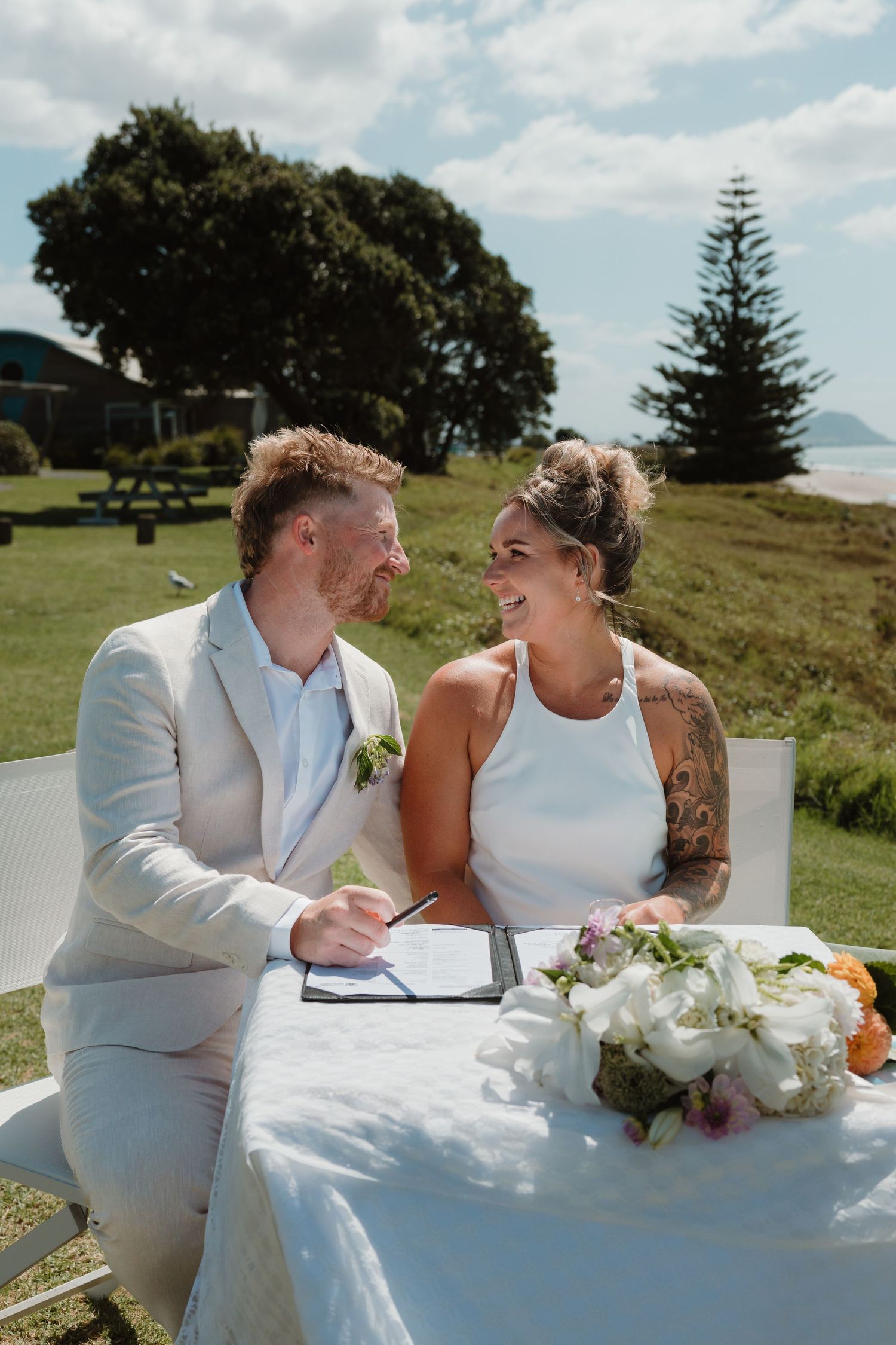 Bride and groom signing wedding register outdoors, smiling at each other, with ocean views and lush greenery behind them.