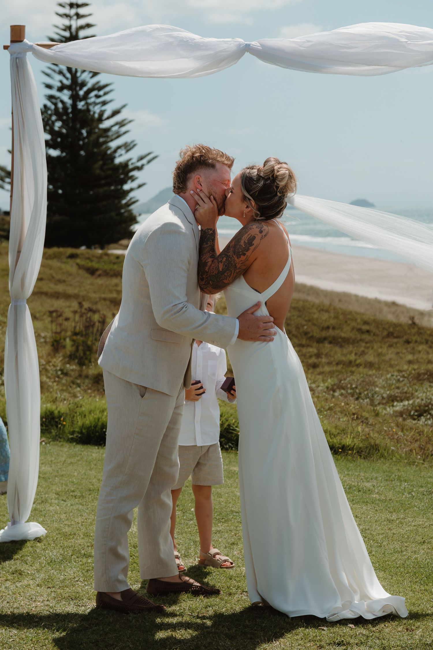 Couple sharing first kiss at outdoor beach wedding ceremony under white draped arch on grassy cliff overlooking ocean.