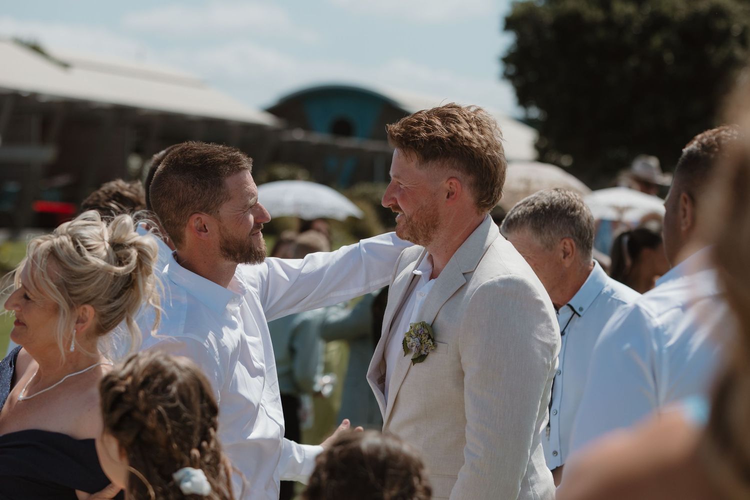 Two men in suits share a warm moment at an outdoor wedding celebration on a sunny day.