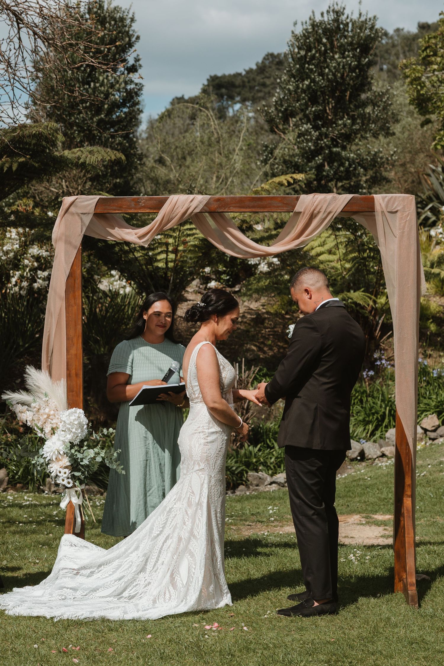 Wedding ceremony at a garden venue with wooden arch draped in blush fabric and floral accents.