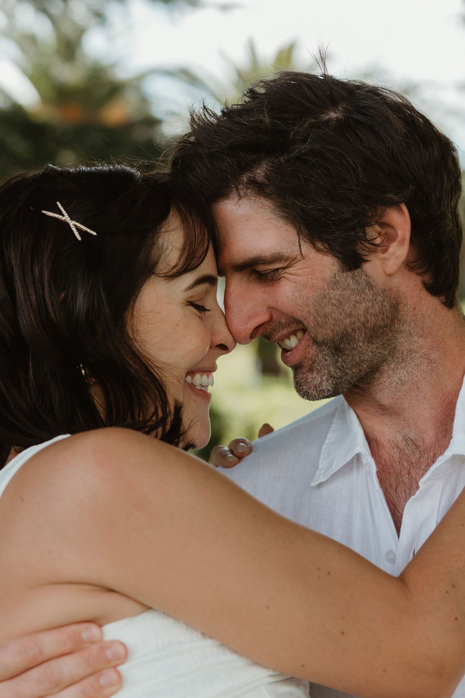 A joyful couple shares an intimate nose-to-nose moment, smiling warmly, with the woman wearing a hair clip outdoors.