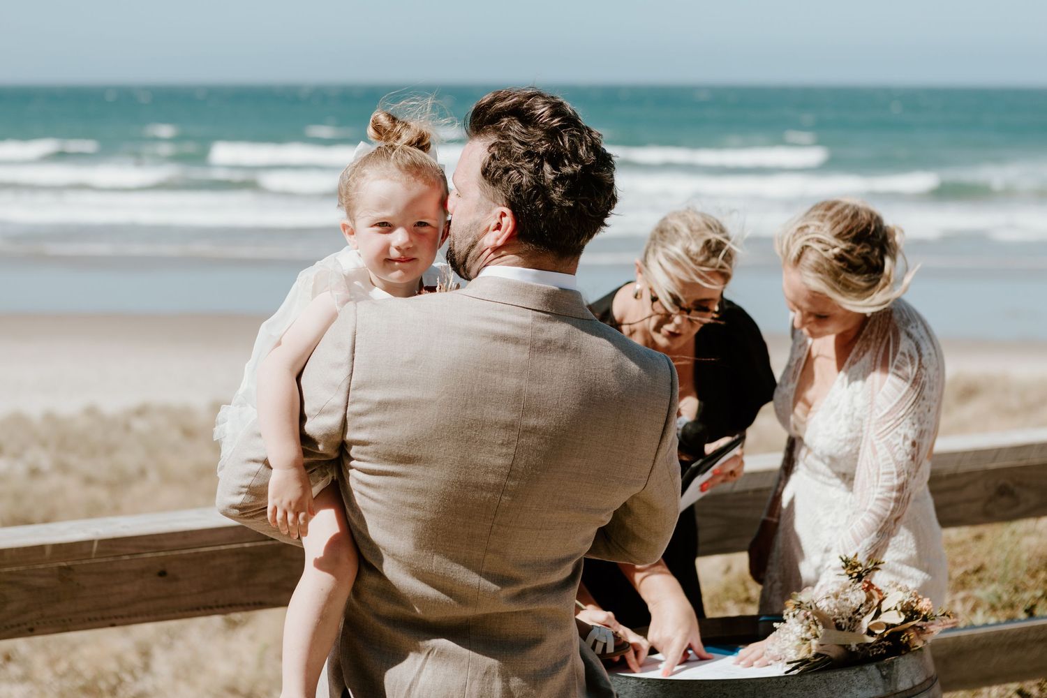 A family enjoying a sunny day together at the beach, sitting by a wooden railing with waves in the background.