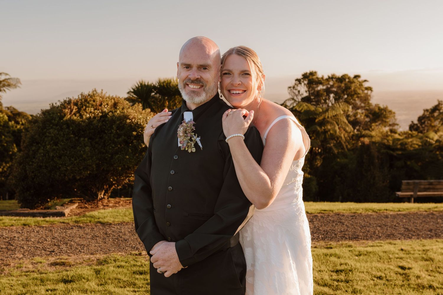 A happy couple embraces in a golden sunlit field during sunset with trees in the background.