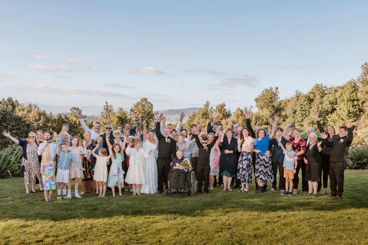 Large wedding party poses together on grassy hill with raised arms celebrating during outdoor summer ceremony.