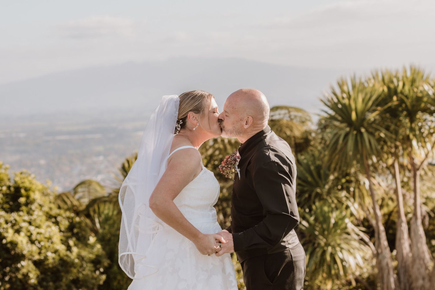 A couple shares a romantic moment on a hilltop overlooking a scenic valley with palm trees in the background.