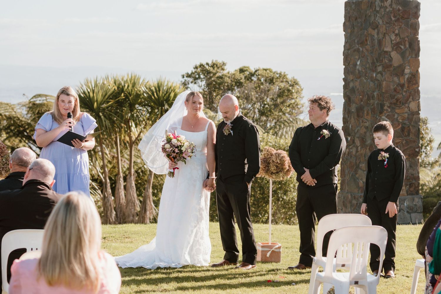 A backyard wedding ceremony scene with palm trees and guests gathered around the couple exchanging their vows.