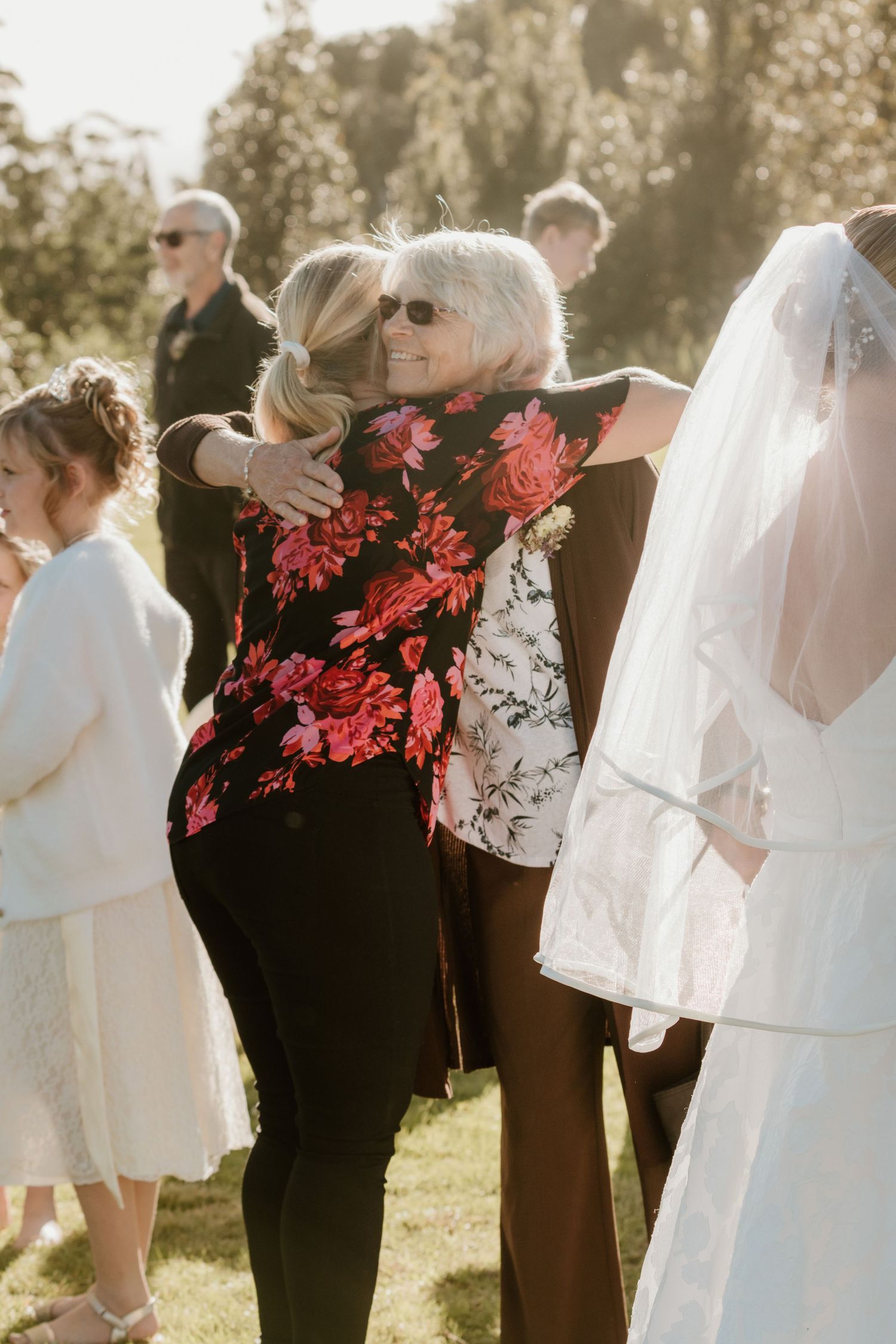 A wedding guest in a black floral dress embraces someone at an outdoor ceremony on a sunny afternoon.