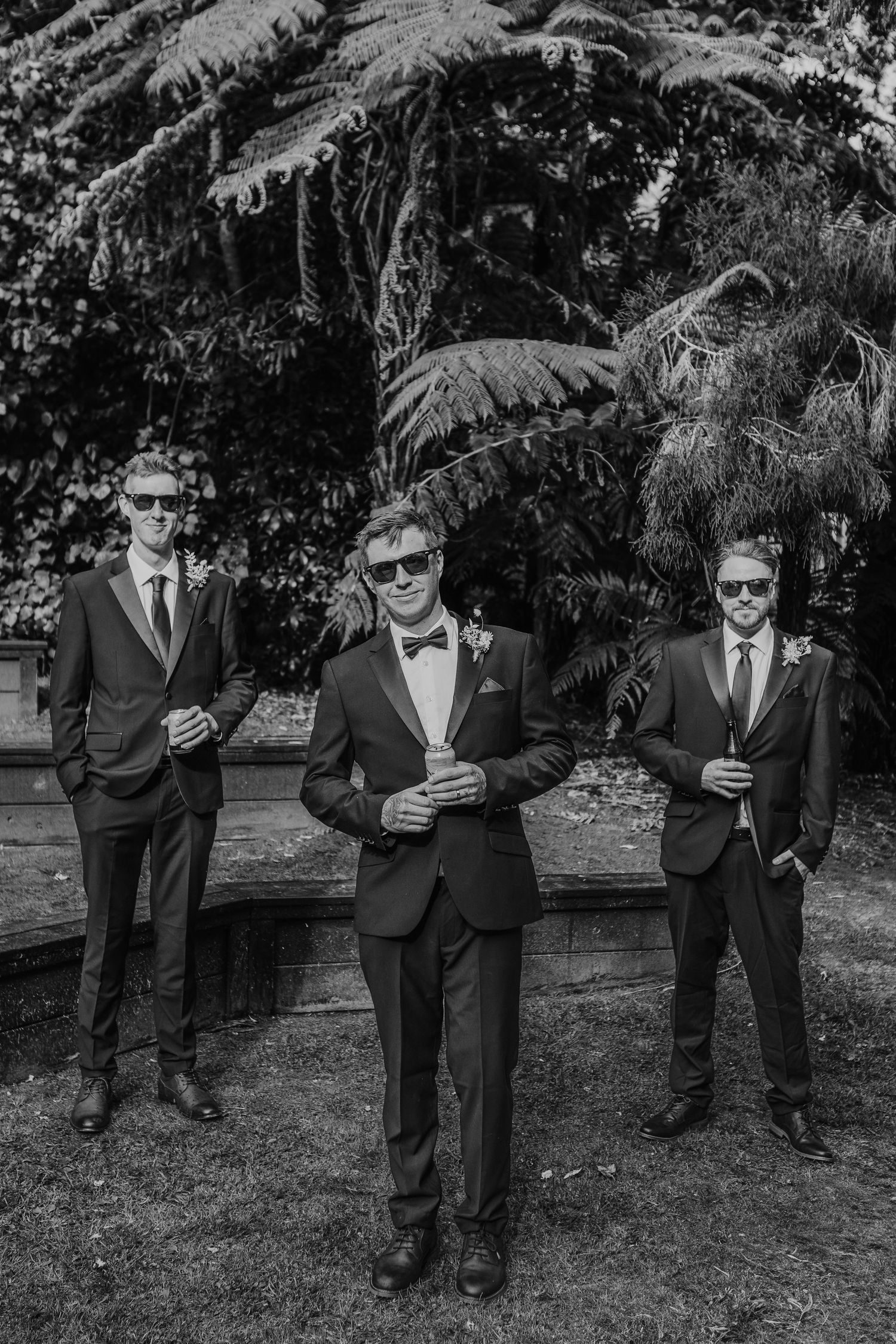 Black and white photograph of three men in formal suits and sunglasses standing on grass with trees in the background.