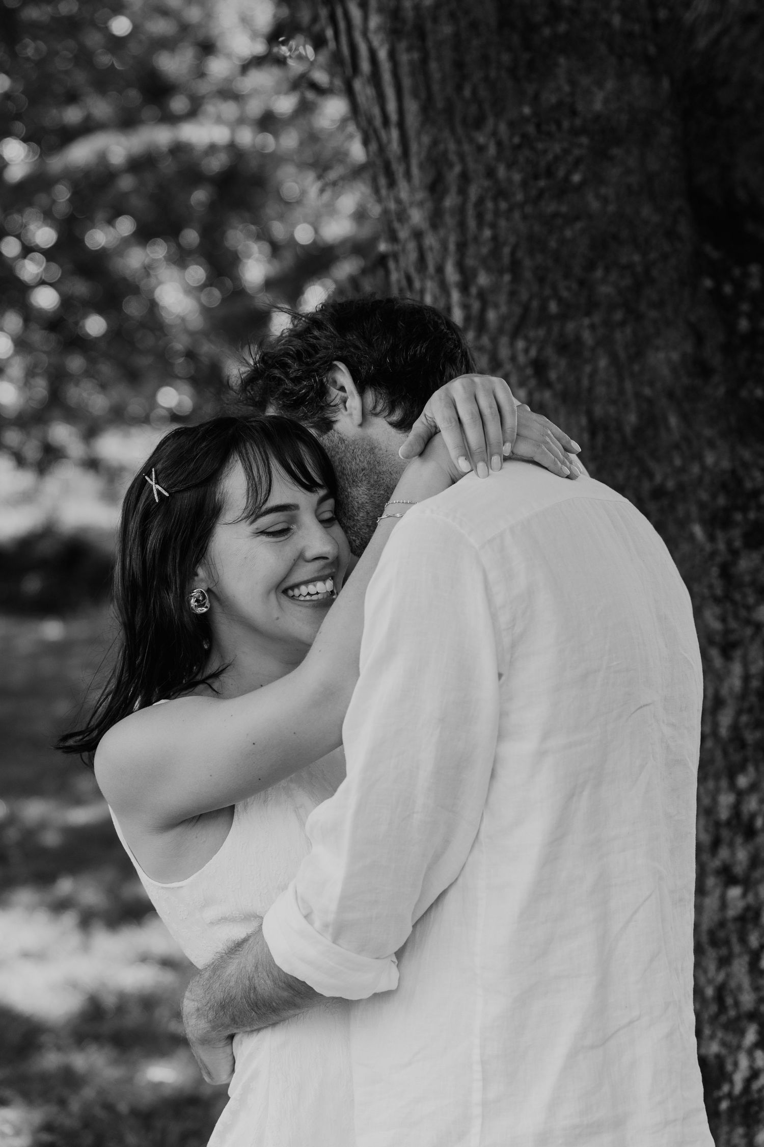 Black and white photo of a smiling woman embracing a man in a white shirt near a tree outdoors.