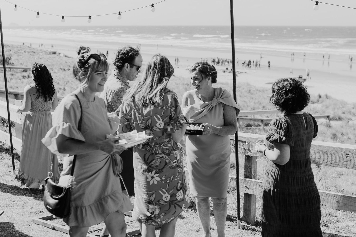 Wedding guests mingle outdoors under string lights at a casual beach reception. Papamoa Surf Lifesaving Club Wedding