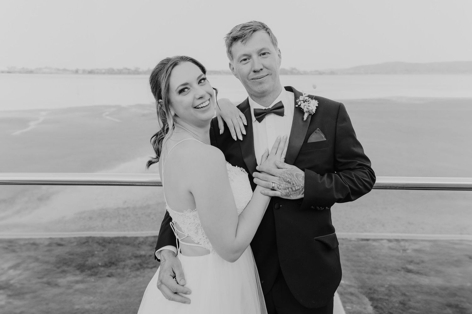 Bride and groom in formal wedding attire posing together with waterfront view in background.