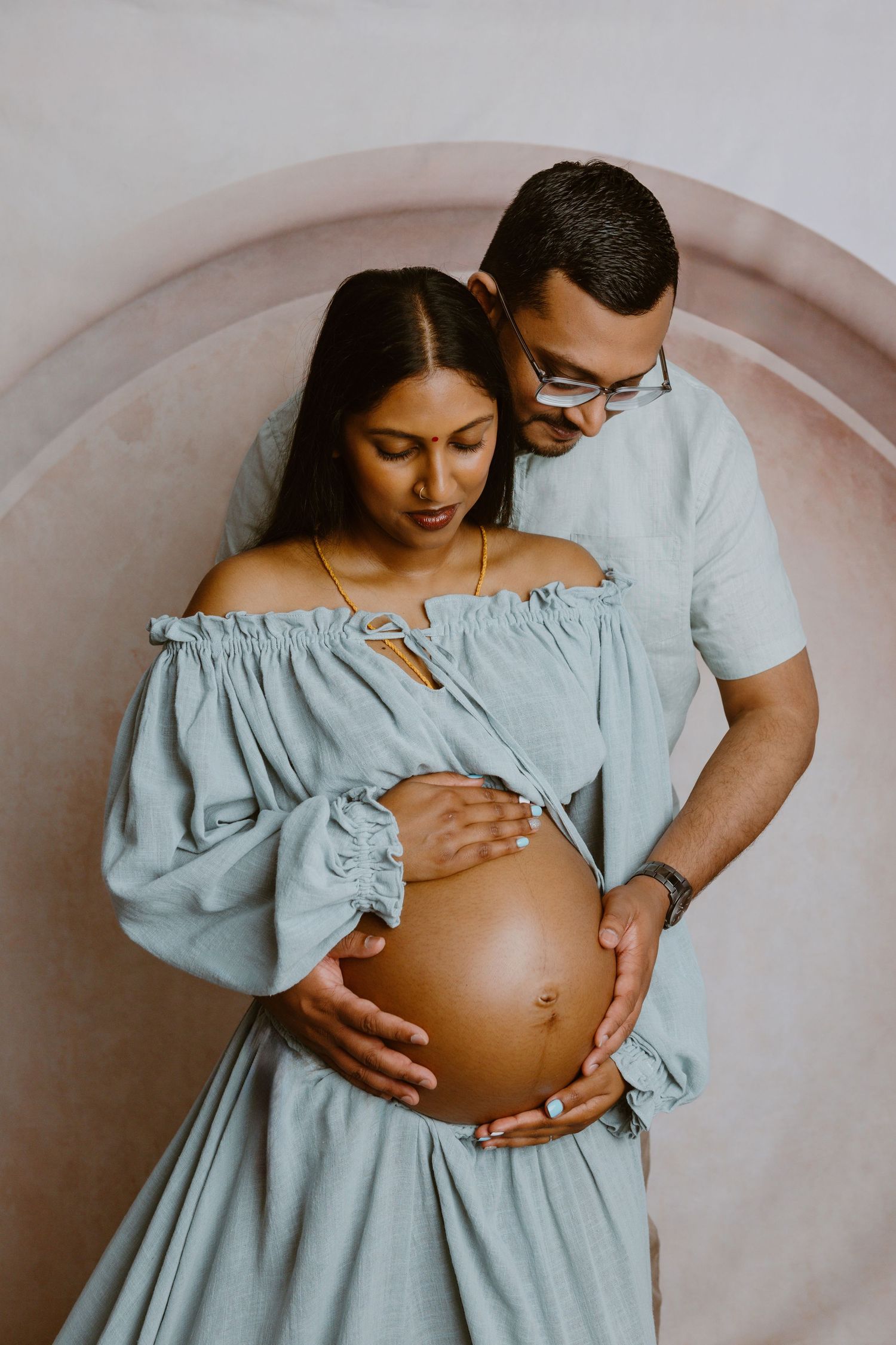 An expectant couple shares an intimate maternity portrait against a neutral backdrop, both wearing soft blue tones. Tauranga maternity