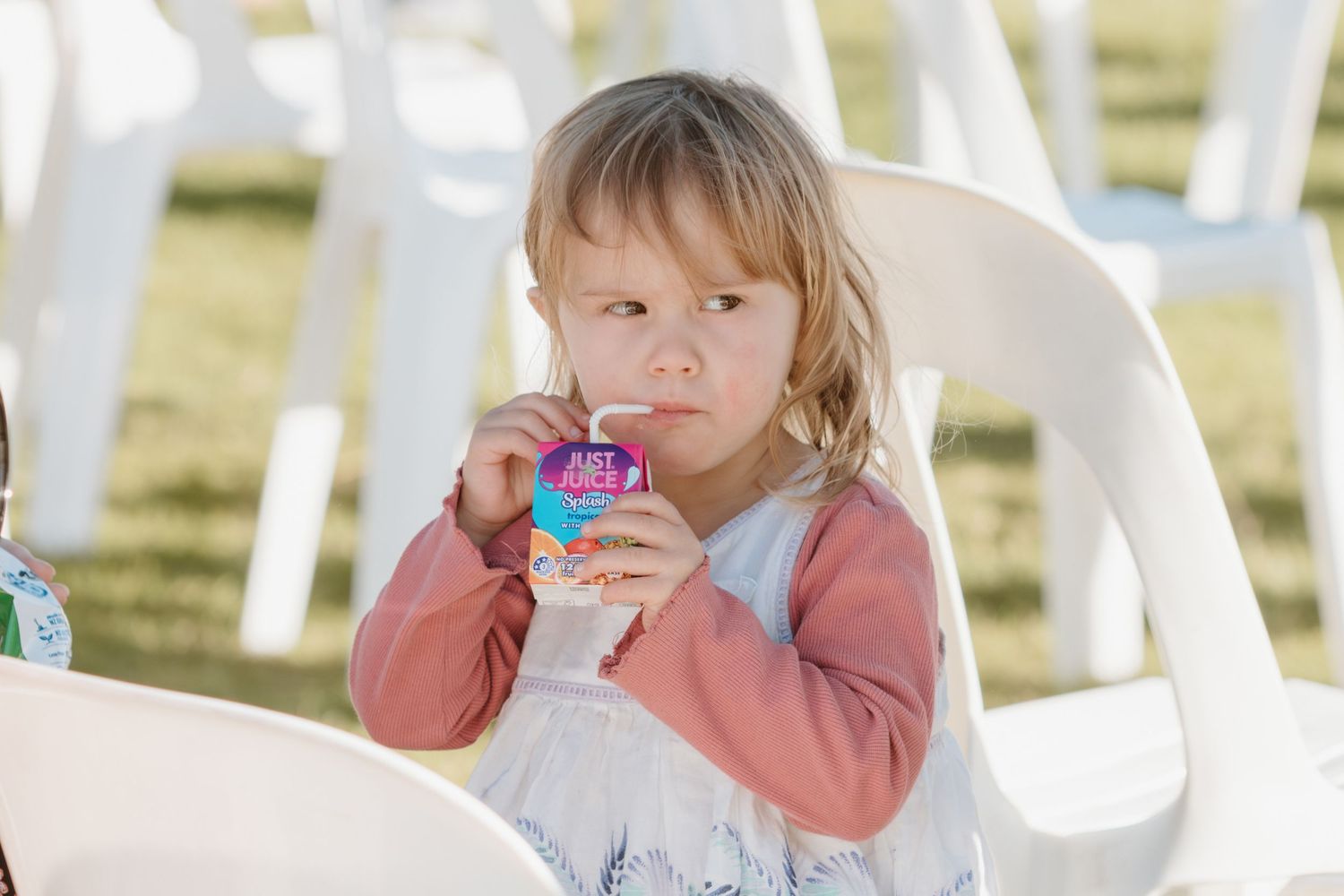 A young child sits in a white outdoor chair drinking from a colorful beverage cup.