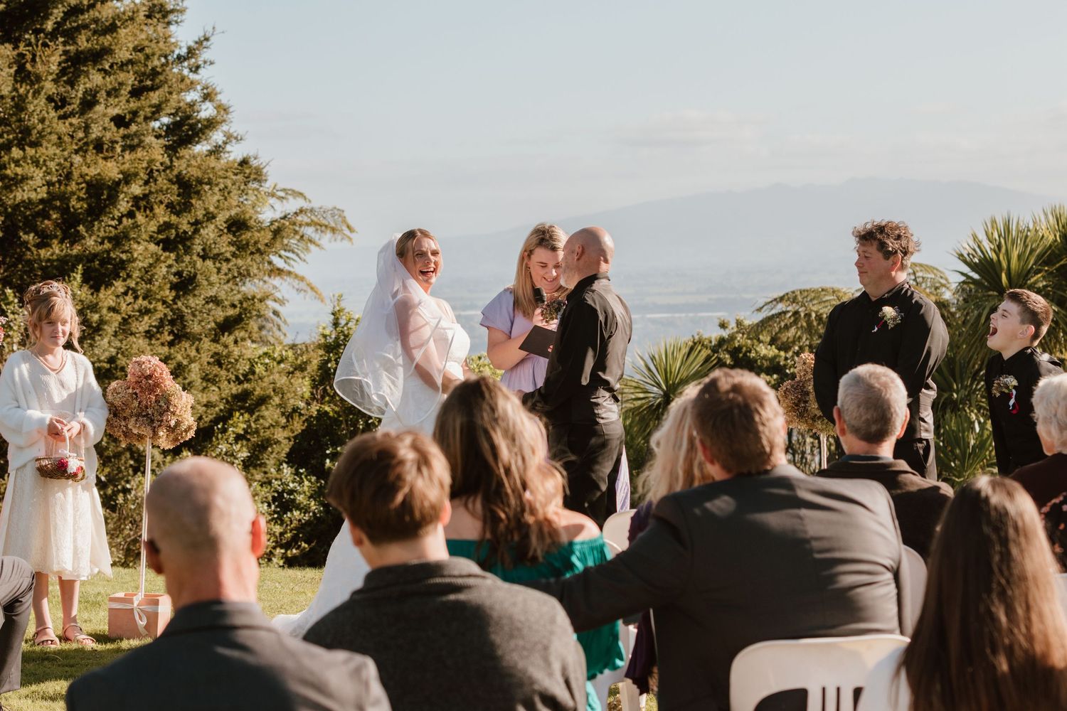 An outdoor wedding ceremony takes place on a hilltop with scenic views and evergreen trees in the background.