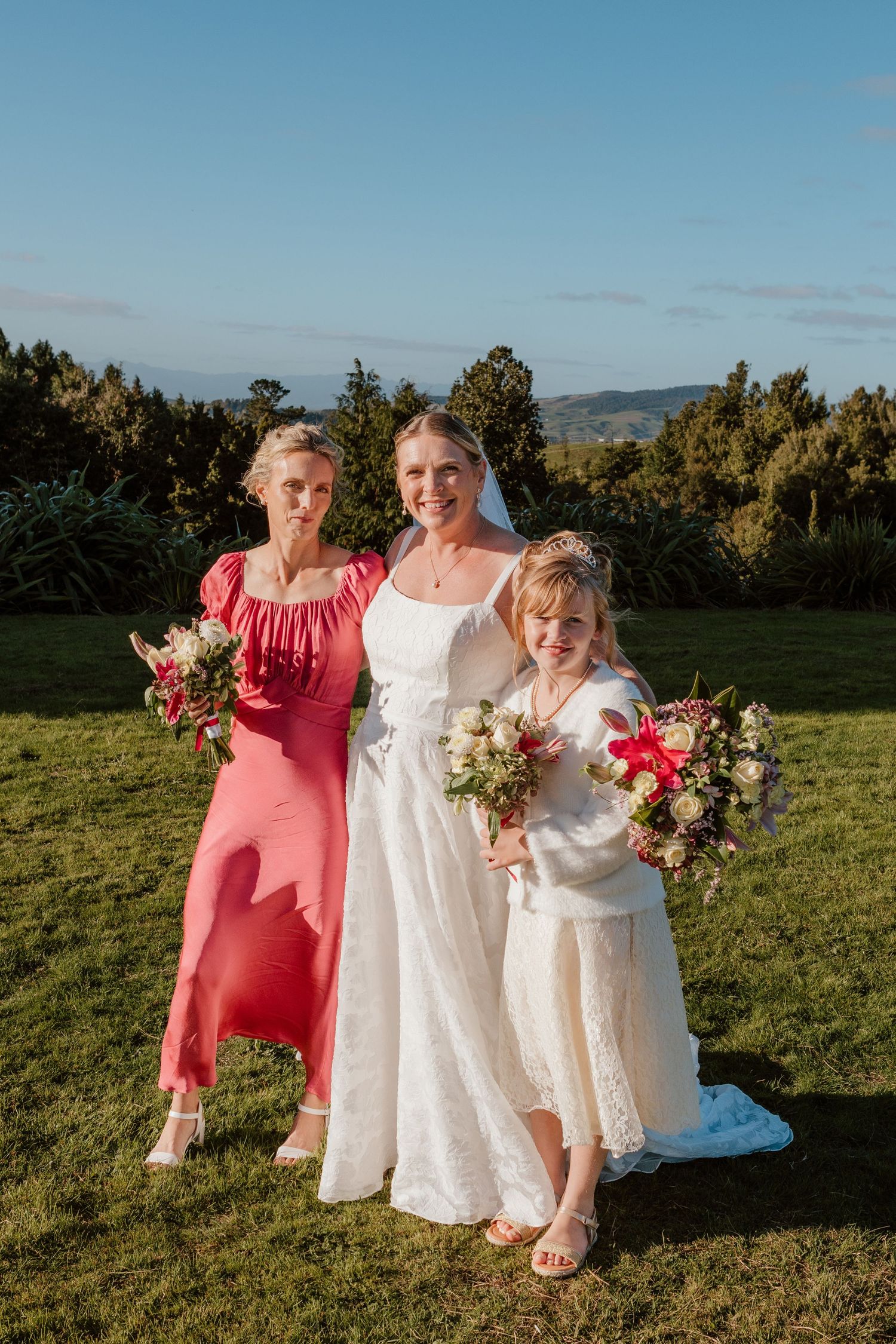 Three figures in white and coral dresses pose together on a grassy hillside at sunset with bouquets of flowers.