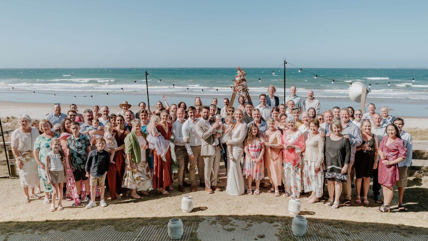Wedding guests gathered for panoramic beach photos with ocean waves and sandy shore in the background.