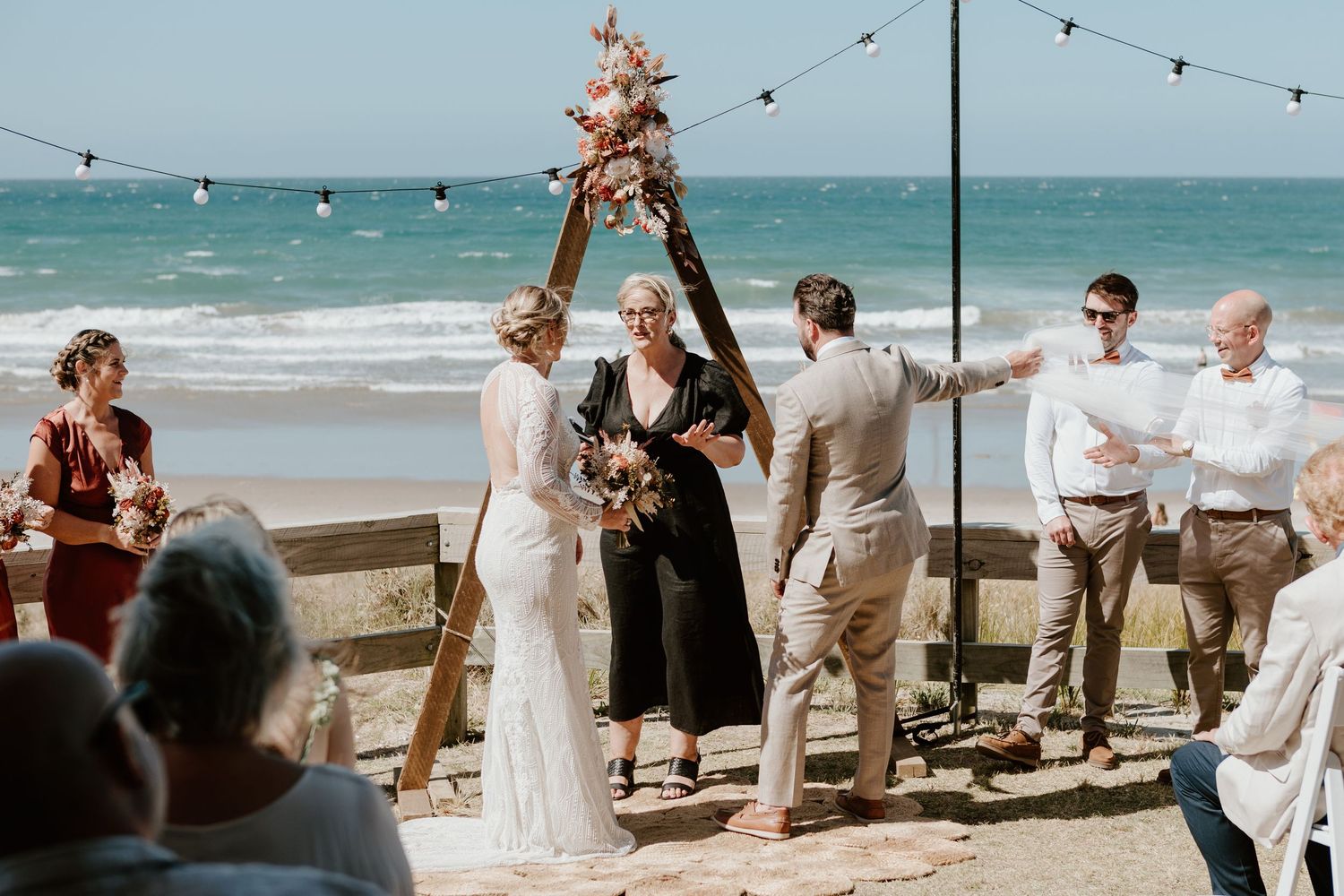 A beach wedding ceremony takes place under string lights with the ocean as a backdrop on a sunny afternoon.