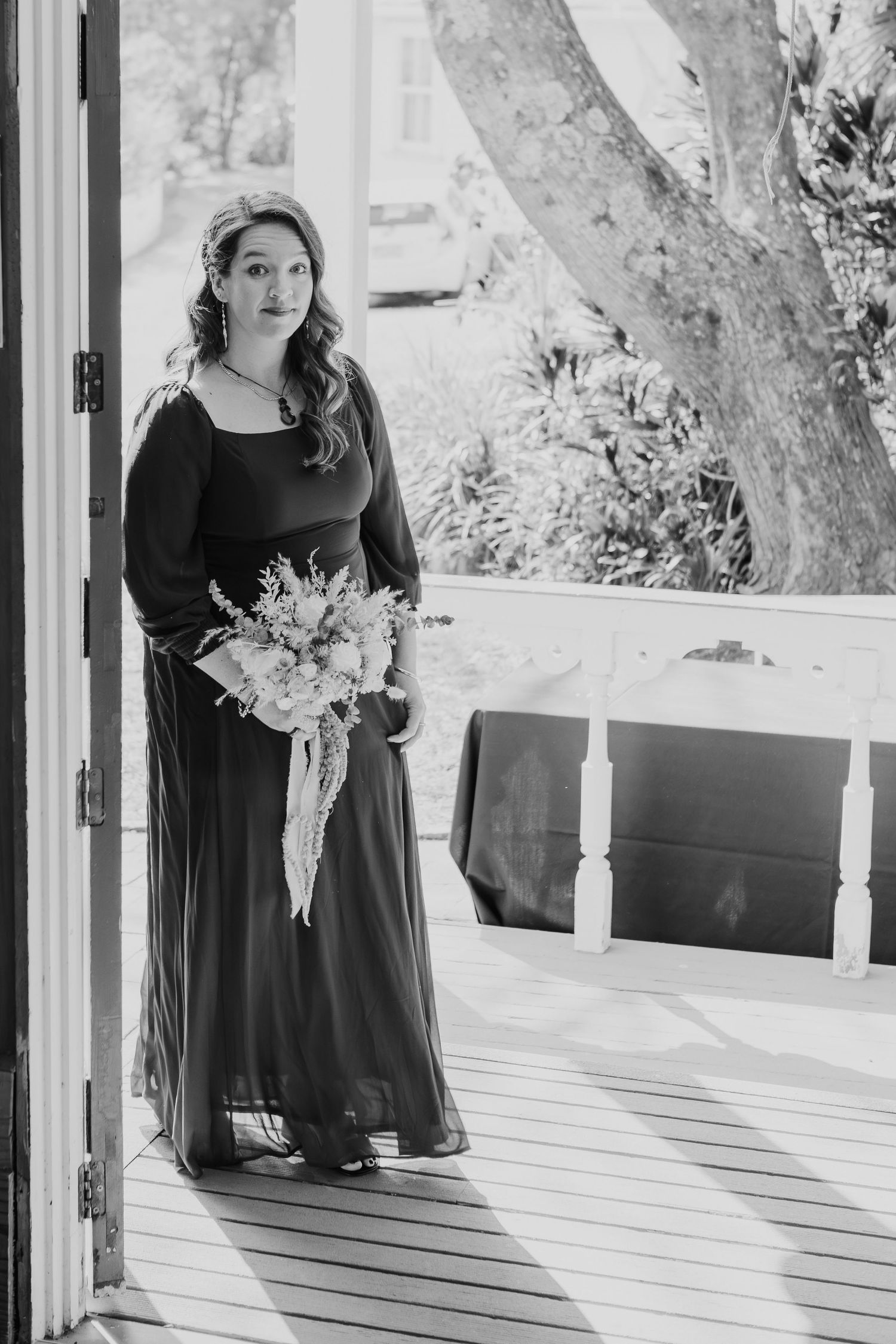 A bride in a black dress holds a floral bouquet while standing on a wooden deck in black and white photography.
