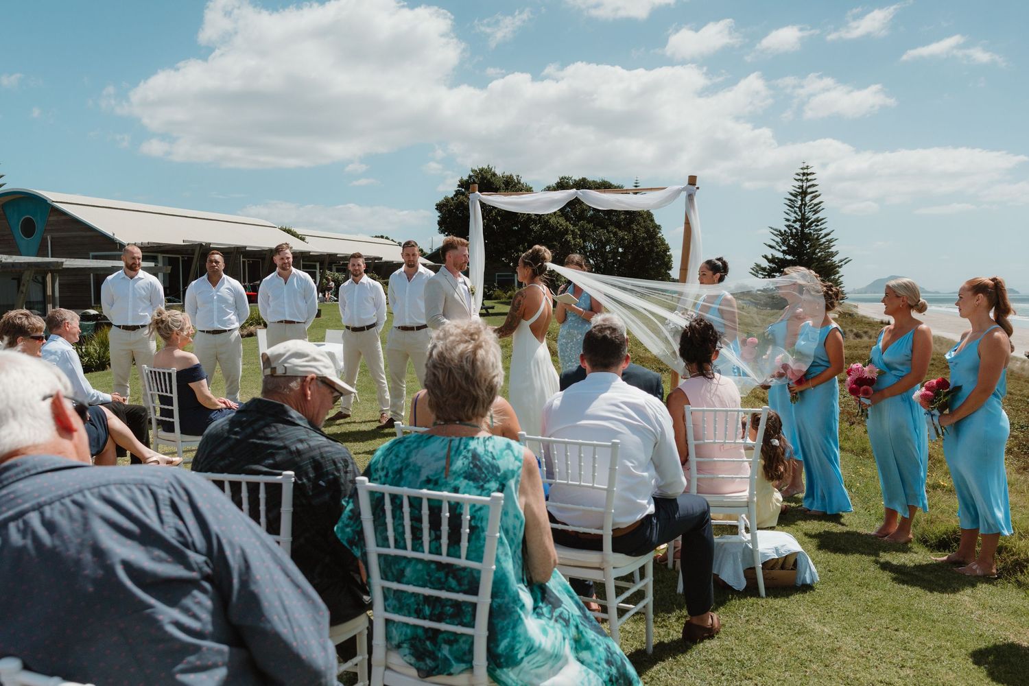 Outdoor beach wedding ceremony with bridesmaids in blue dresses, groomsmen in white, and guests seated on white chairs.