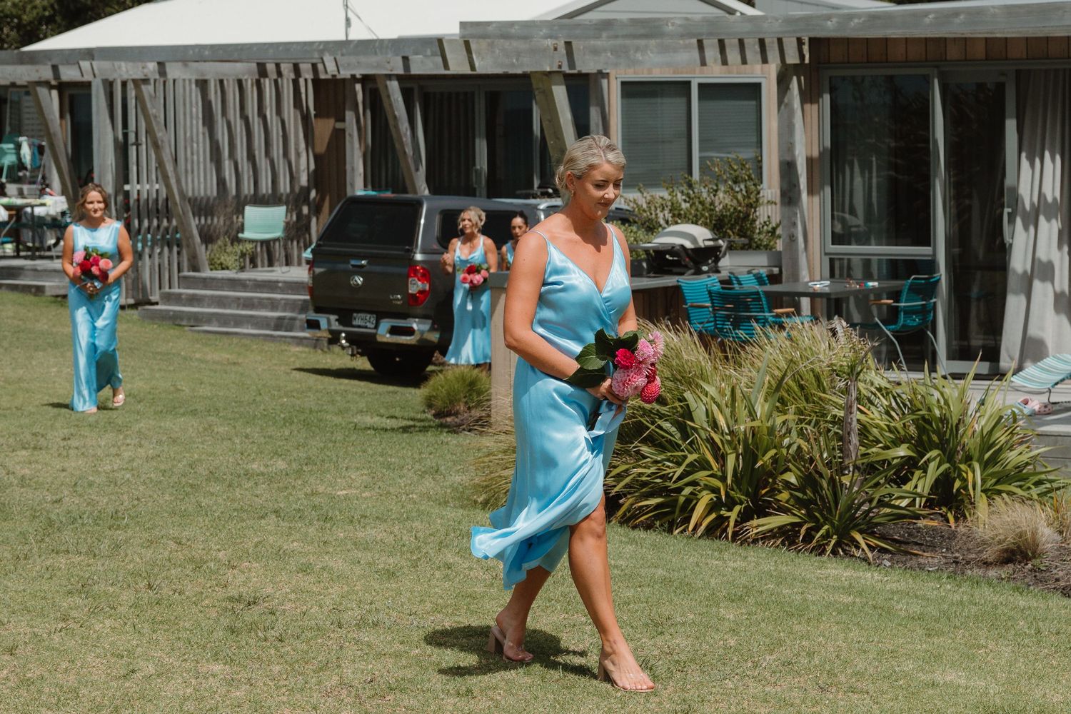 Bridesmaids in turquoise dresses walking across green lawn at outdoor wedding ceremony on sunny day.