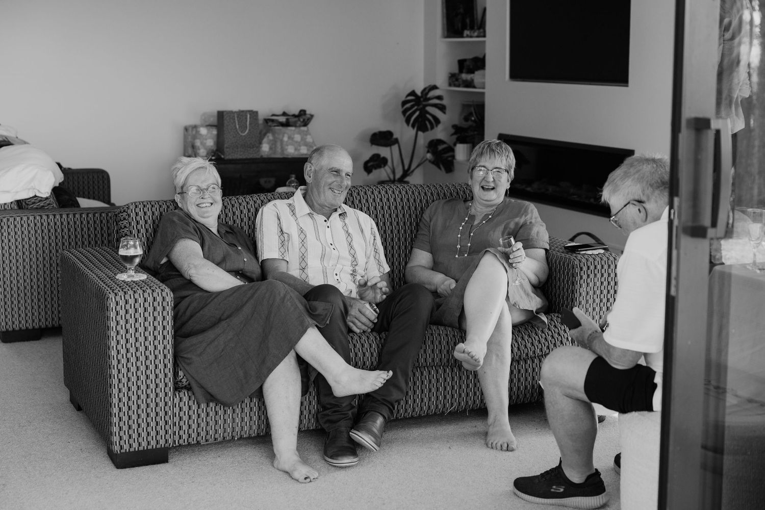 Black and white photo of four elderly people sitting and laughing together on a sofa in a cozy living room.
