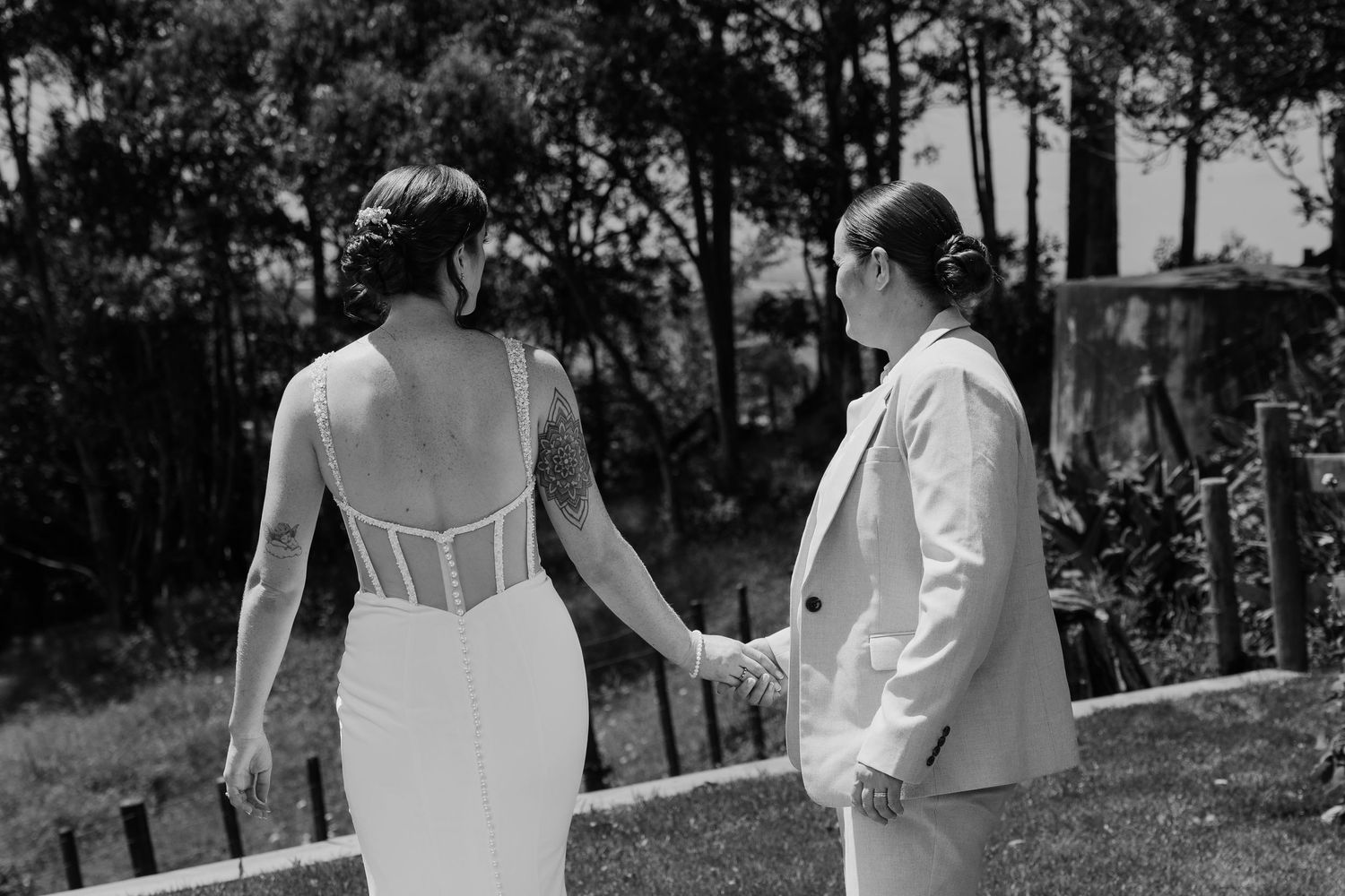 Black and white photo of two brides holding hands, one in a backless wedding gown with tattoos, the other in a suit.