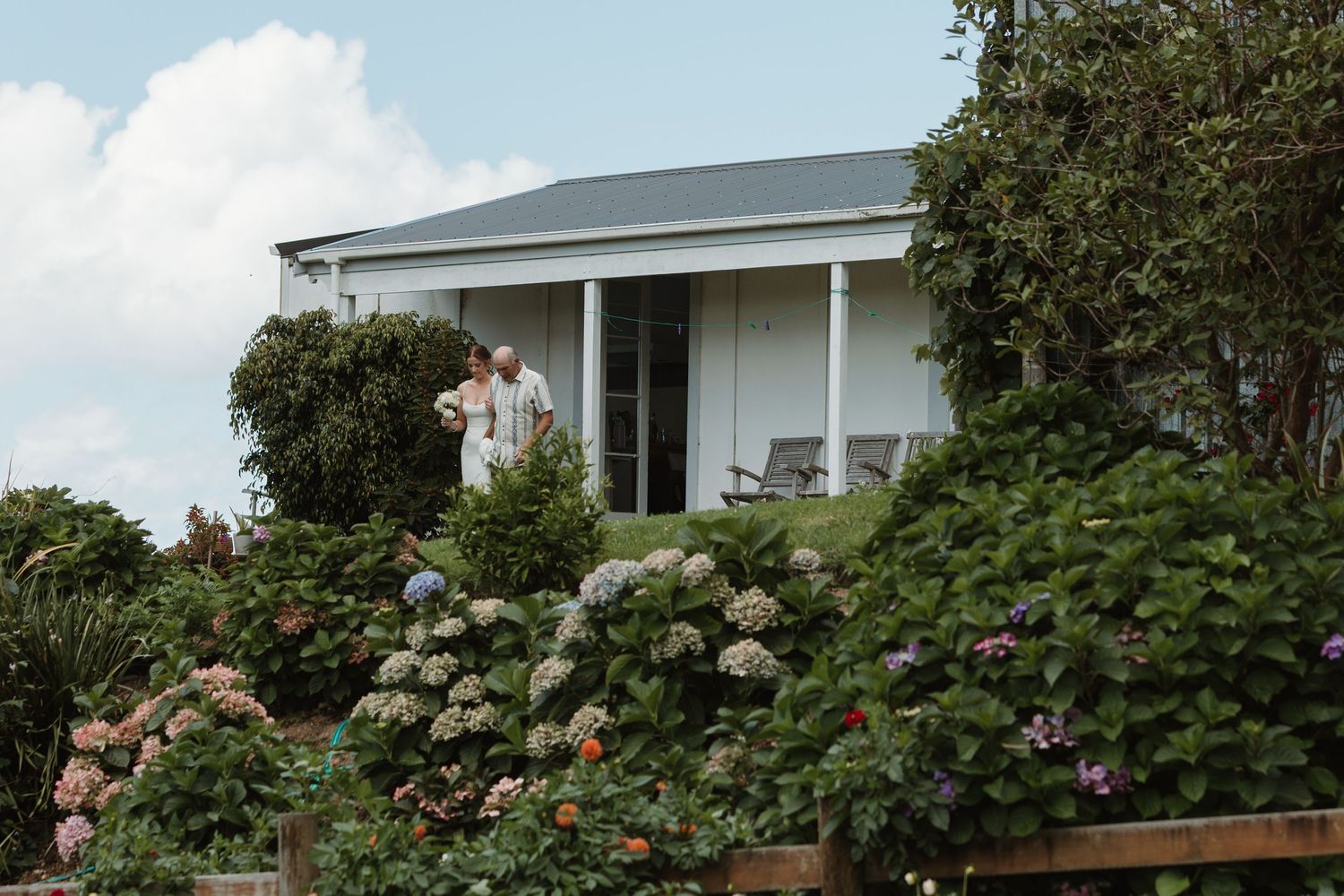 Couple embracing on a white cottage porch surrounded by lush hydrangea gardens under a cloudy sky.