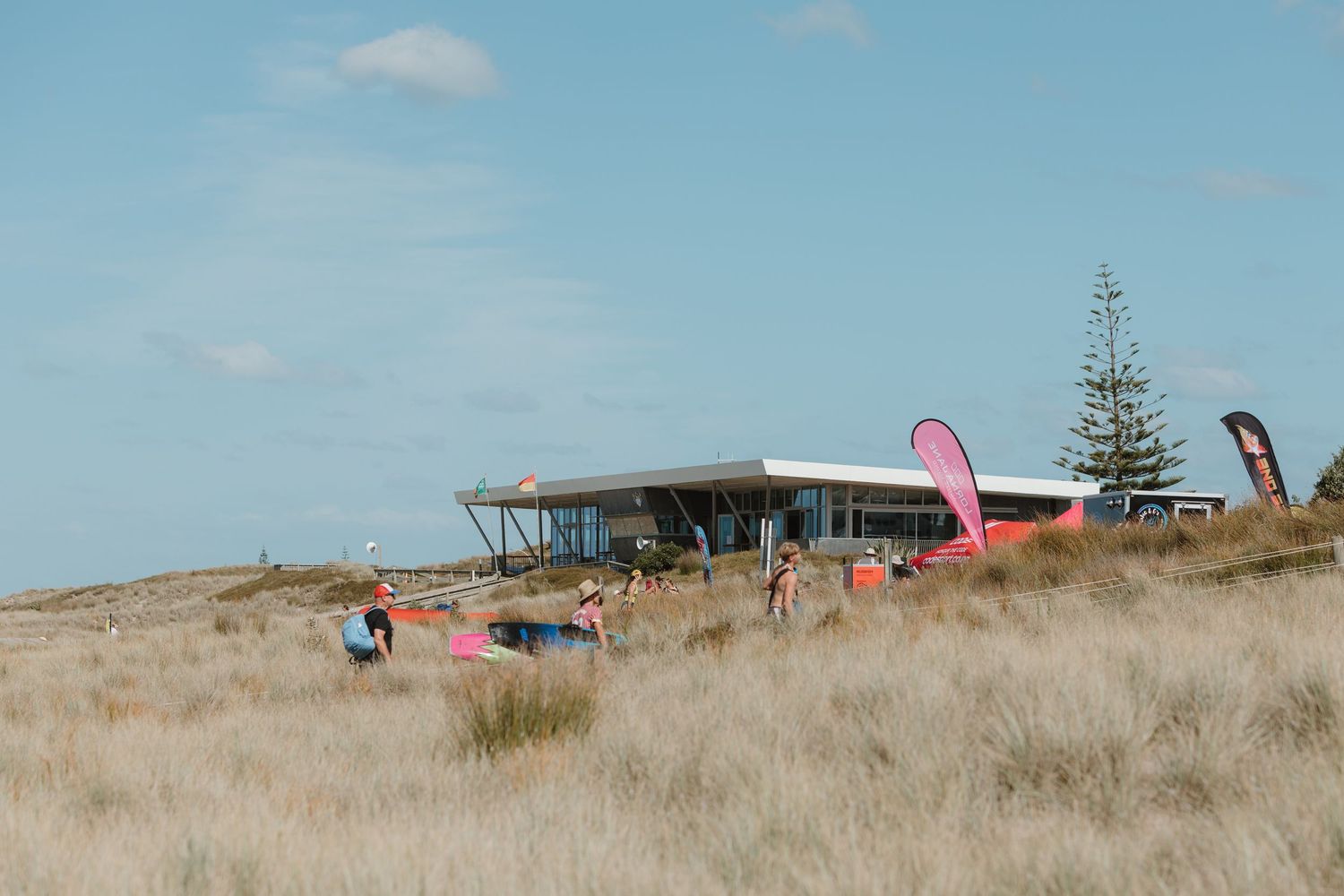 Surfers and beachgoers gather near a modern coastal clubhouse surrounded by golden dune grass under a clear blue sky.