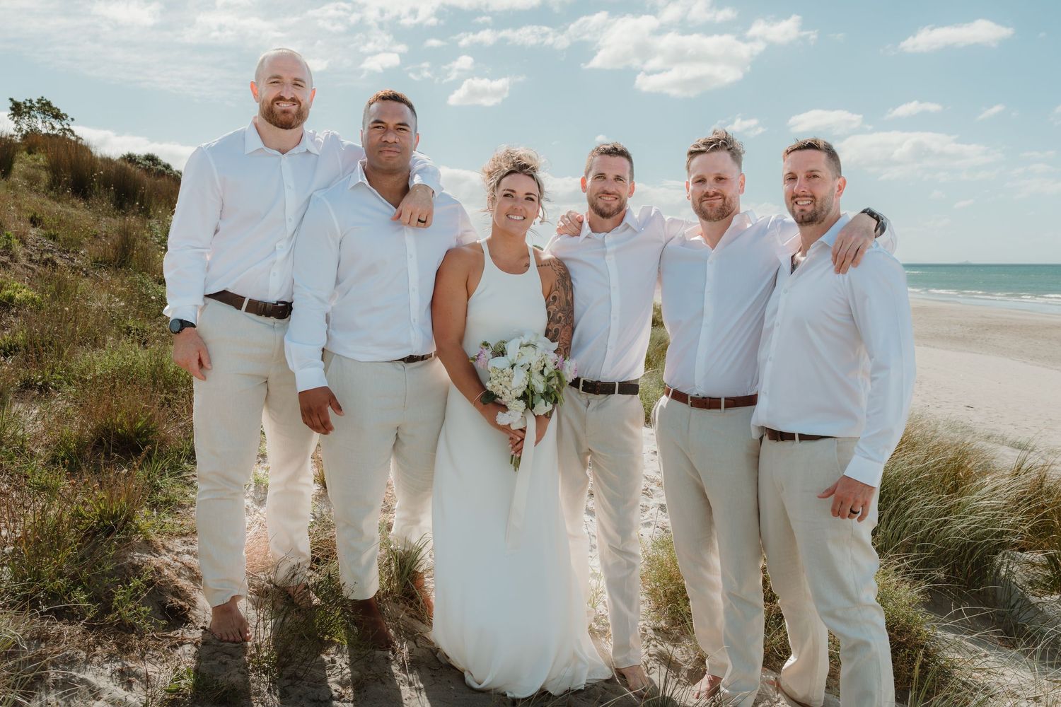 Bride and groomsmen posing together on a sandy beach, wearing white shirts and tan pants at an outdoor wedding.