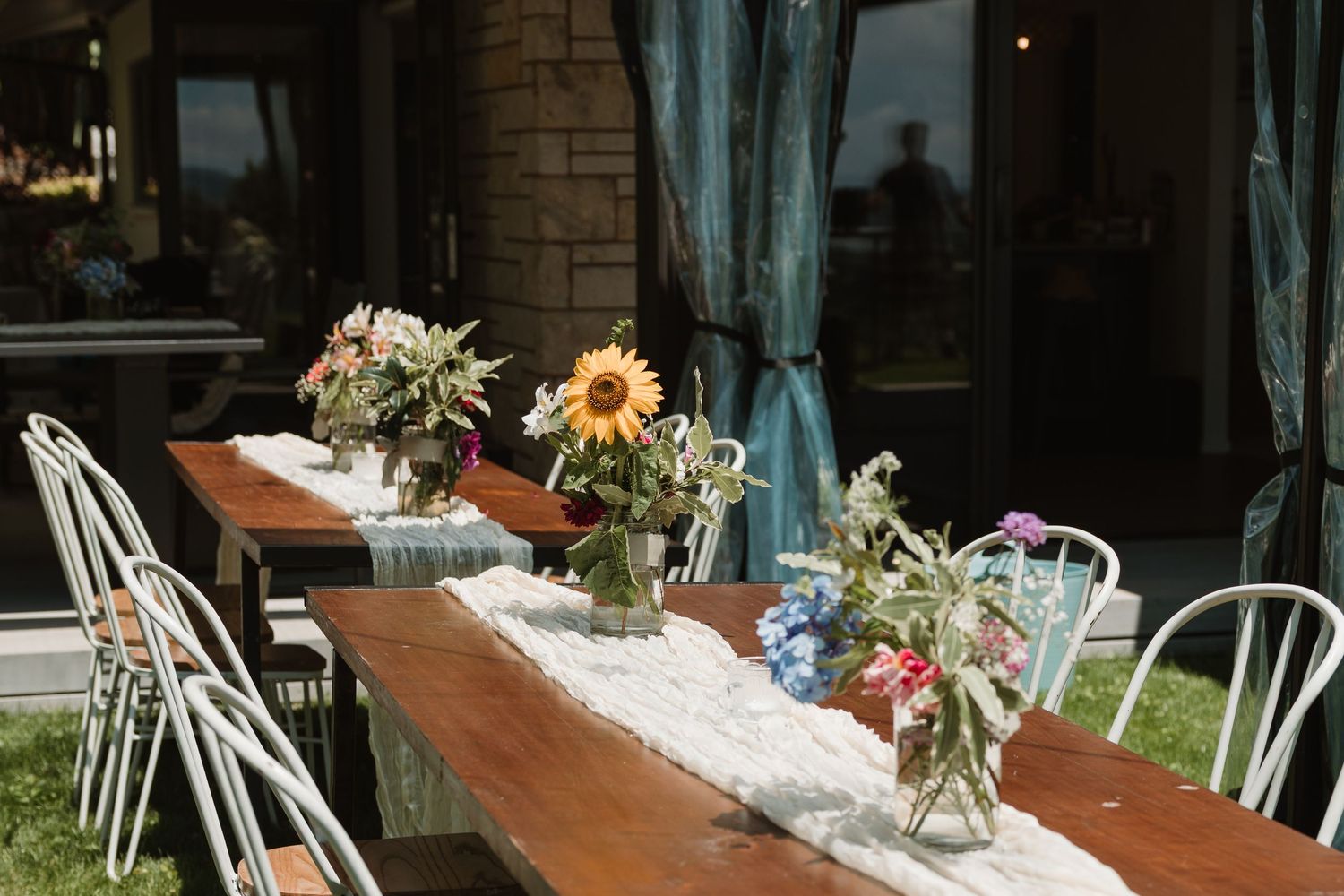 Rustic wooden outdoor dining table with floral centerpieces including sunflowers, surrounded by white chairs on a sunny day.