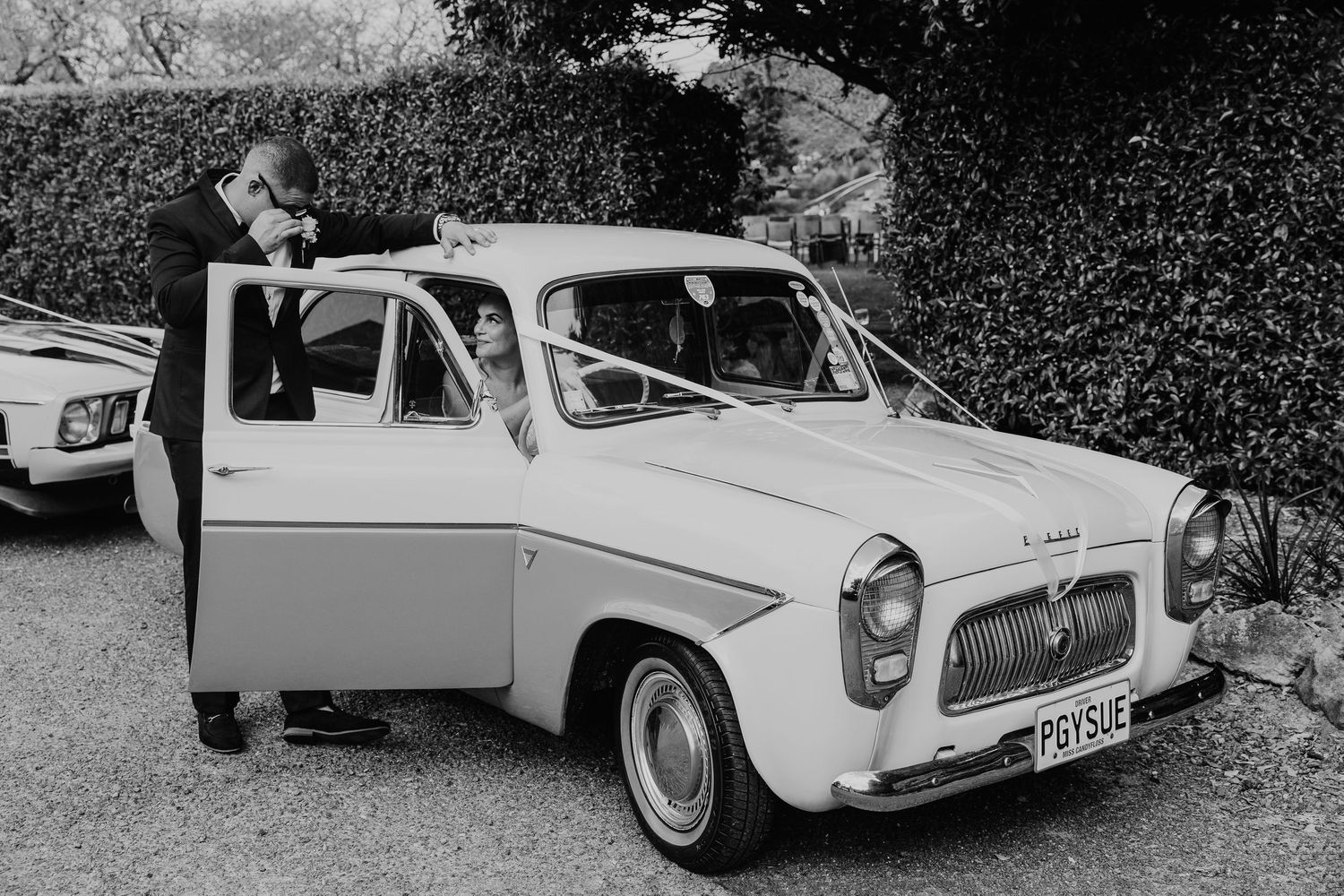 Vintage black and white photo of a Ford Anglia 100E car from the 1950s with open doors parked near hedges.