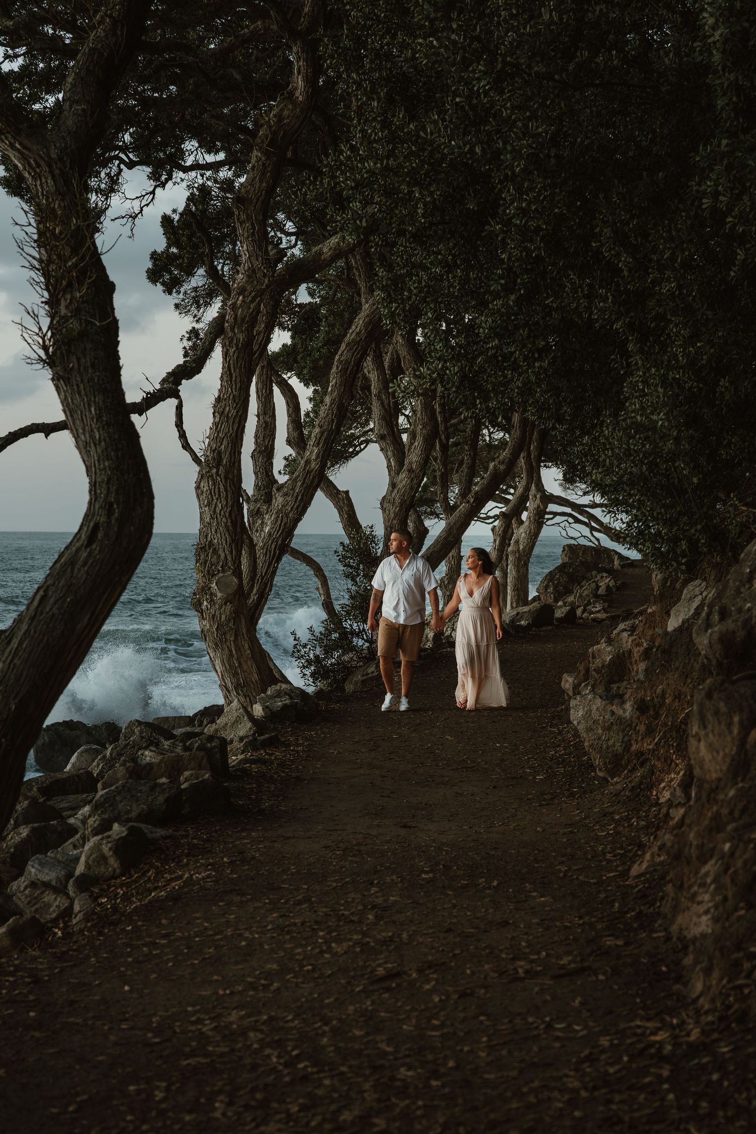 A scenic coastal path under twisted trees with people walking along the ocean's edge at sunset.