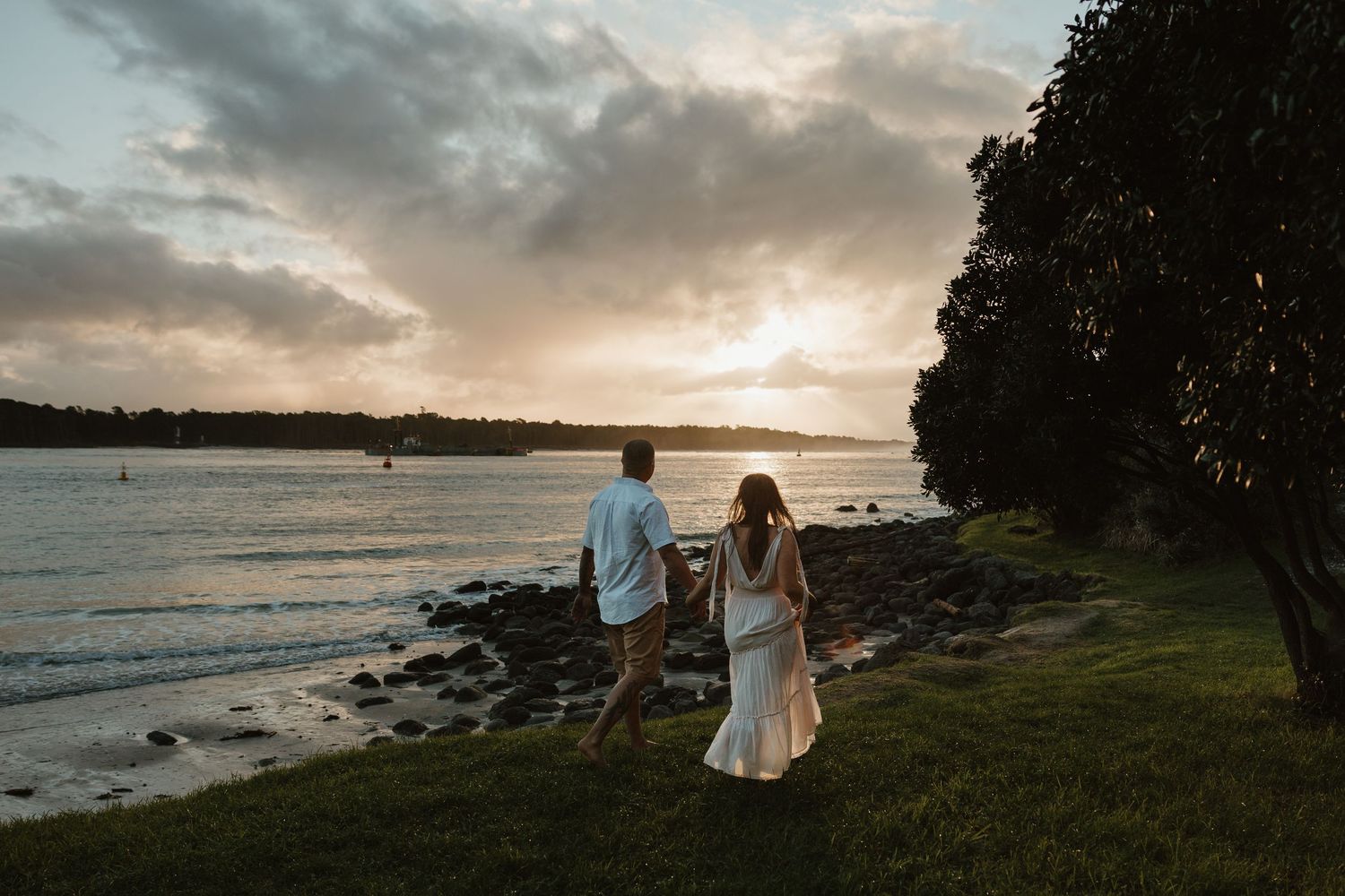 A couple walks along a scenic coastal path at sunset as waves crash against the shore in the background.