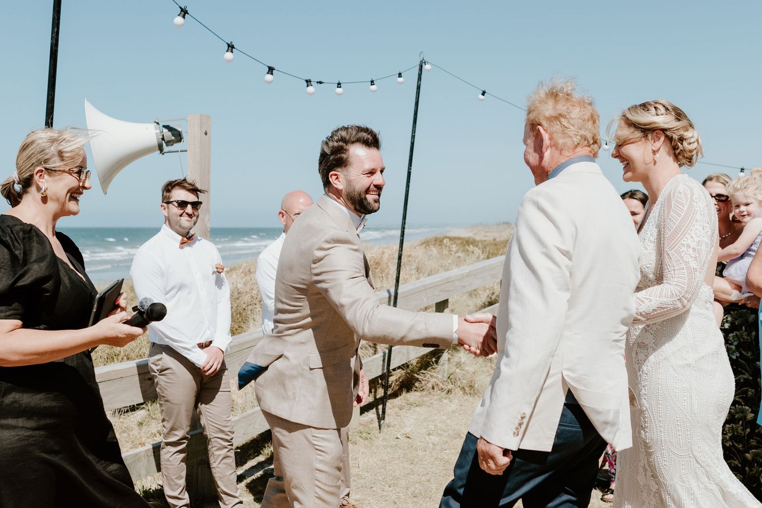 Beach wedding celebration with string lights and guests in light-colored attire against blue sky. Papamoa Surf Lifesaving Club Wedding